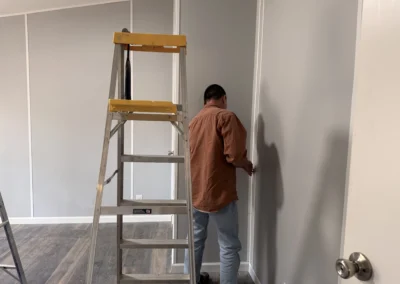 A worker in a brown shirt and jeans standing next to a silver step ladder against a gray paneled wall